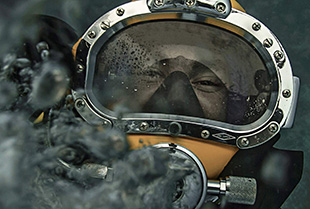 A United States Navy diver poses for a picture under water in full diving gear.